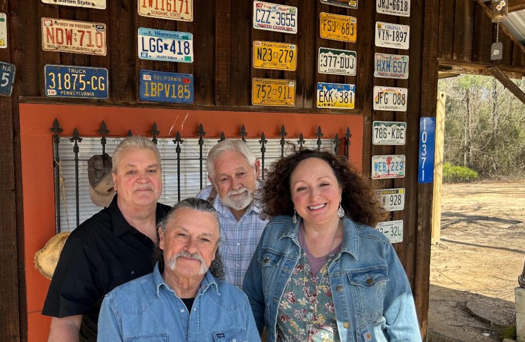 Four smiling adults, three men and one woman, pose in front of a rustic wooden building decorated with vintage license plates. They are standing near a window with iron bars and a collection of hanging hats.