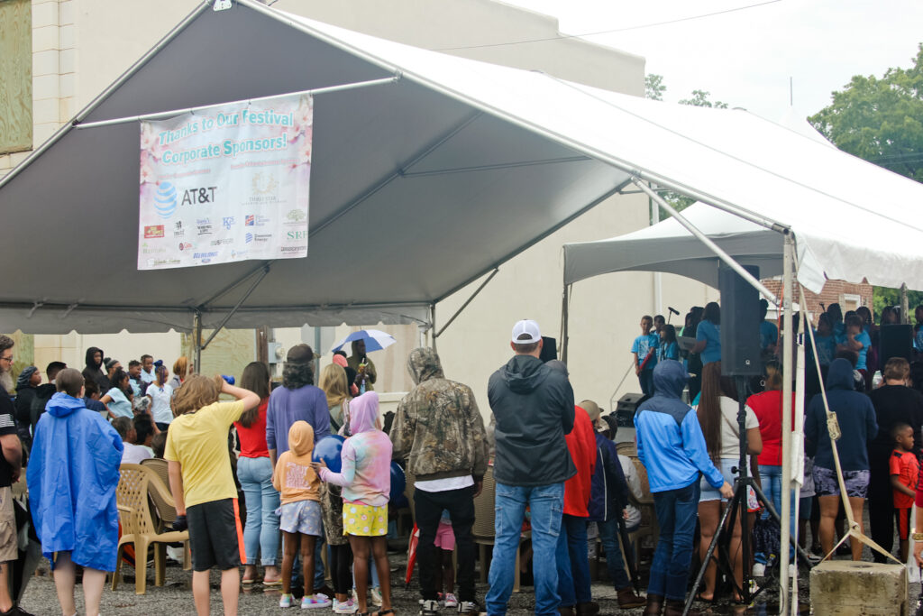 A crowd of people, including children and adults, gather under and around a large white canopy tent at an outdoor event. A group of children performs on a stage to the right, while a banner above thanks corporate sponsors.