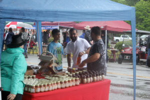 A group of people interacting under a blue canopy at an outdoor market on a rainy day. A table in the foreground is covered with jars and bottles of sauces or condiments. Tents, shoppers with umbrellas, and a red utility vehicle are visible in the background.