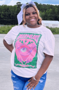 A smiling woman with blond-highlighted hair and a white bow wears a white T-shirt with a colorful graphic and ripped jeans, standing outdoors on a concrete surface.