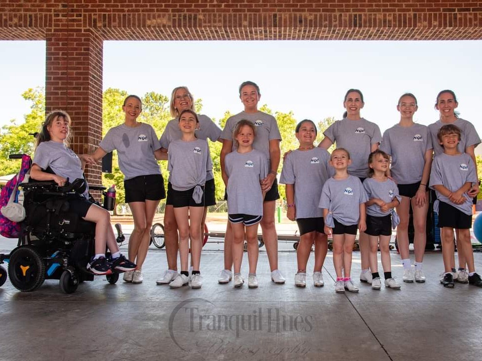 A diverse group of children and adults, including a participant in a wheelchair, stand in a line wearing matching gray 'NGC' t-shirts and black shorts. They are smiling and posing under a pavilion with brick columns.